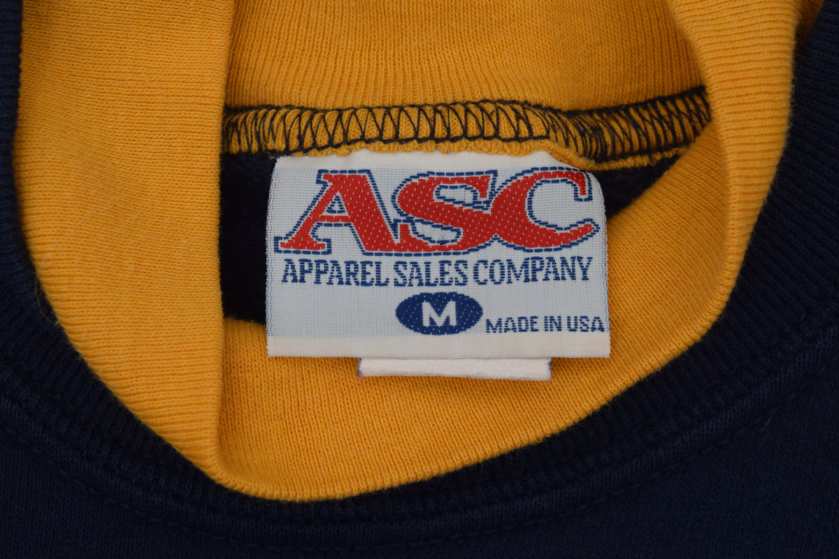 Close-up of the inside collar of a University of Michigan crewneck sweatshirt showing a white sewn-in label for ASC Apparel Sales Company with red and blue lettering, set against a yellow ribbed collar and navy blue knit fabric.