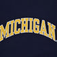 A close-up of a navy blue University of Michigan crewneck sweatshirt featuring the embroidered word 'MICHIGAN' with yellow varsity-style lettering with a white outline.