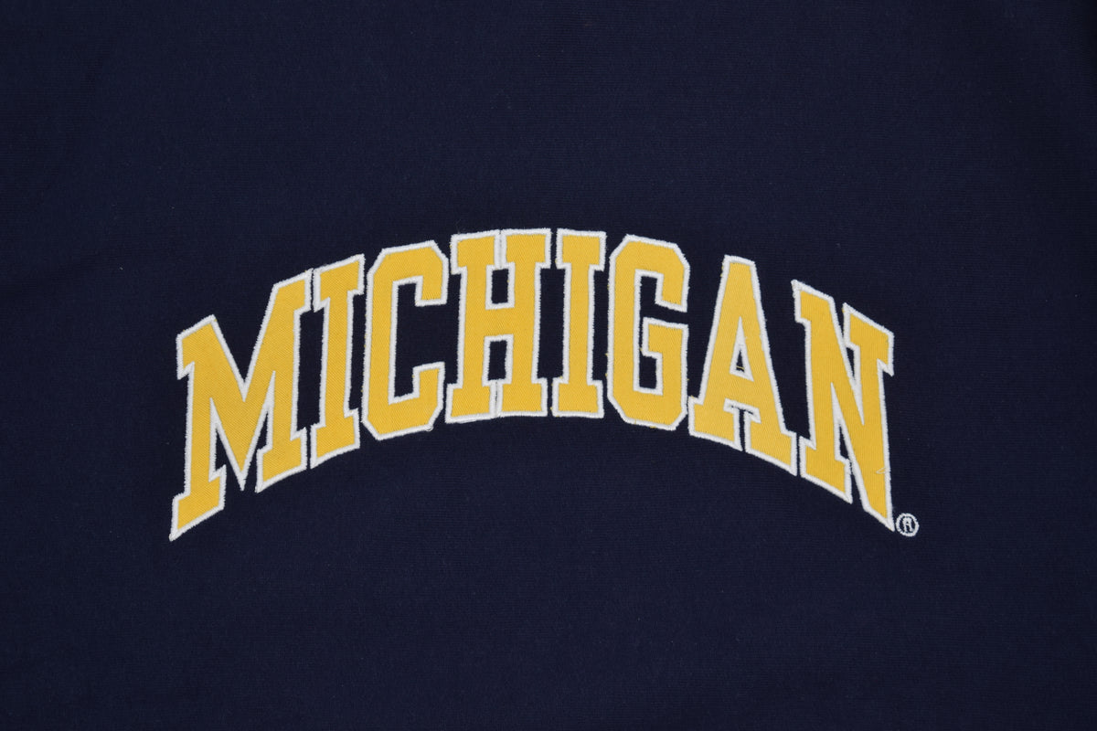 A close-up of a navy blue University of Michigan crewneck sweatshirt featuring the embroidered word 'MICHIGAN' with yellow varsity-style lettering with a white outline.