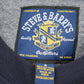 Close-up of the inside neck label of a University of Michigan navy blue crewneck sweatshirt, showing the brand 'Steve & Barry's Outfitters' embroidered on a blue patch with a gold crest.