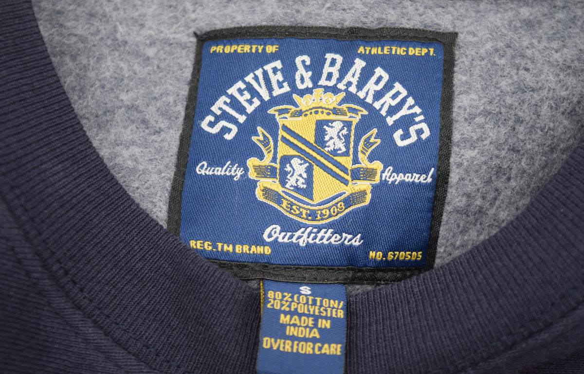 Close-up of the inside neck label of a University of Michigan navy blue crewneck sweatshirt, showing the brand 'Steve & Barry's Outfitters' embroidered on a blue patch with a gold crest.