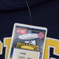 Close-up of a University of Michigan navy blue sweatshirt with yellow and white lettering, featuring a hanging tag for Steve & Barry's.